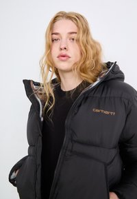 Svart puffjakke med glidelås, hette og elastiske mansjetter. Har en lettvektig design og subtil Carhartt-merkevare i gull.