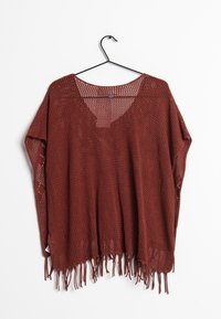 Rostfärgad stickad poncho med fransar längs fållen, vida ärmar och V-ringning, hängande på en svart galge mot en vit bakgrund.