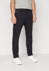 Jeans negros de corte ajustado, hechos de denim. Presenta un detalle de bolsillo con cremallera y una marca sutil. Combinados con zapatillas blancas.