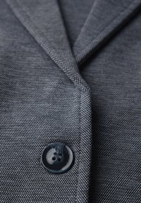 Gros plan sur le devant d'un blazer gris foncé texturé avec un seul bouton noir à quatre trous et des coutures visibles sur le revers.