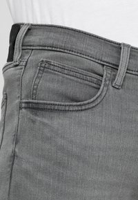 Šedé džíny z denimu s přiléhavým střihem, obsahující styl pěti kapes, viditelné švy a černou nášivku na pase.