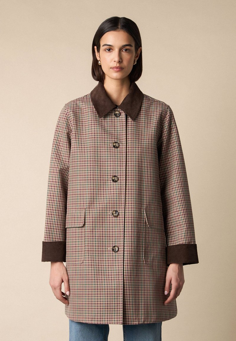 Vrouw draagt een beige en bruin geruite jas van middelhoge lengte met een donkerbruine kraag en manchetten, dichtgeknoopt, staand tegen een effen achtergrond.