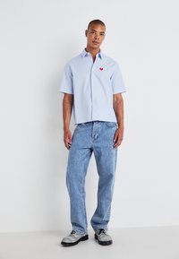 Chemise en jean bleu clair à manches courtes avec un logo en forme de cœur rouge, associée à un jean baggy bleu clair et des chaussures grises avec des lacets noirs.