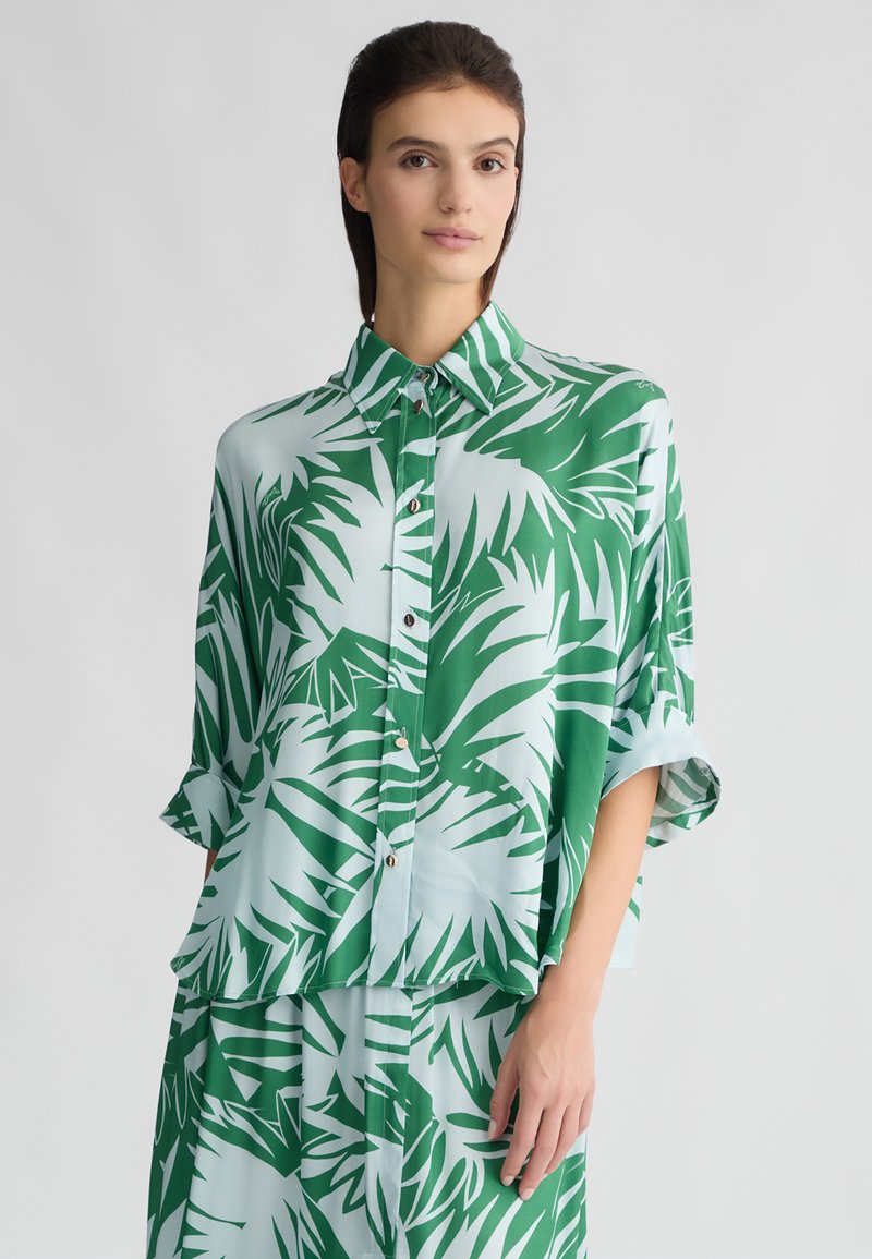 Camicia a maniche corte verde e azzurro chiaro con un motivo di foglie di palma, chiusura con bottoni e vestibilità rilassata. Tessuto liscio e leggero.