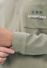 Light green shirt with a textured fabric, featuring a chest pocket. Printed text includes "ASAHIKAWA" and Hokkaido. Fastened with green buttons.