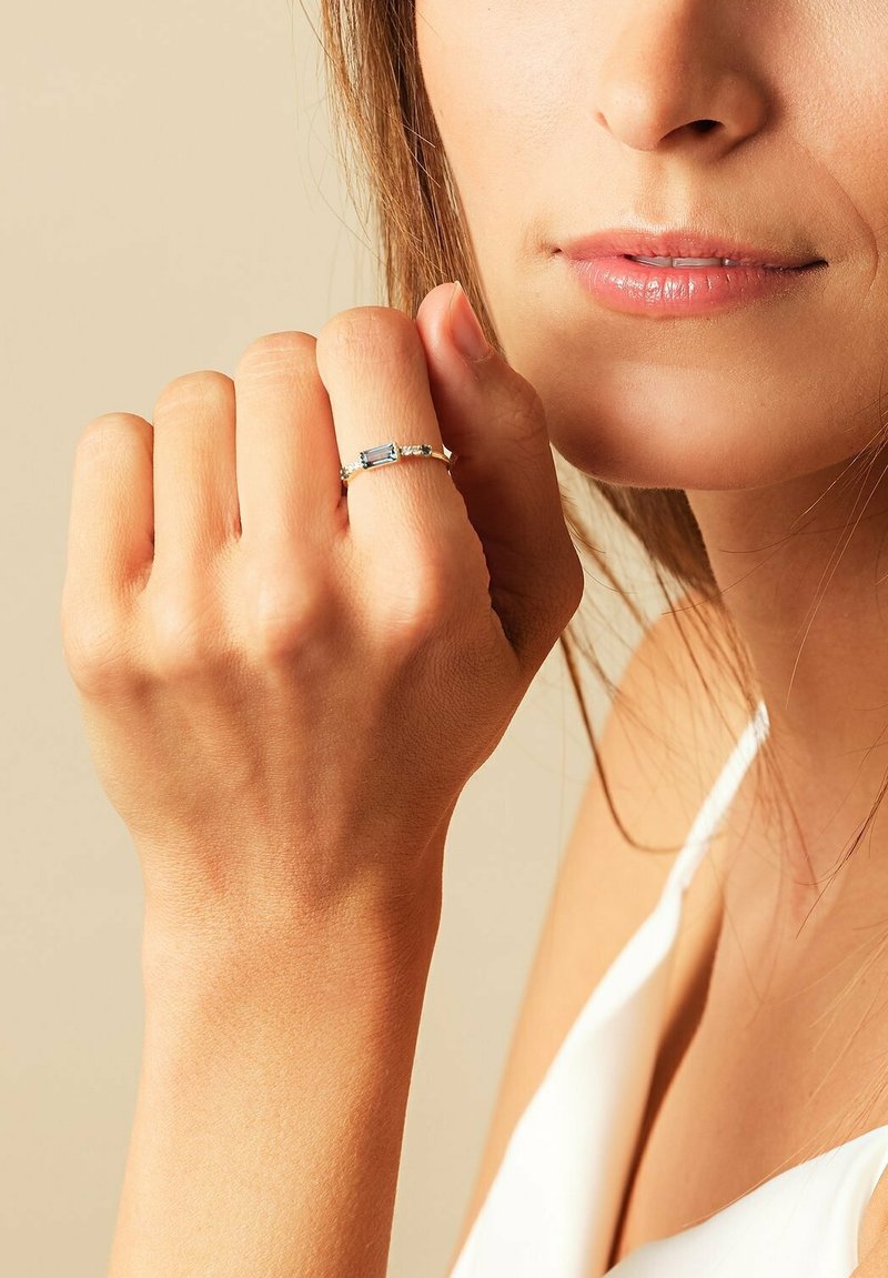 Silberner Ring mit einem rechteckigen blauen Edelstein, filigrane Details im Ringband, getragen an einer Hand vor einem neutralen Hintergrund.