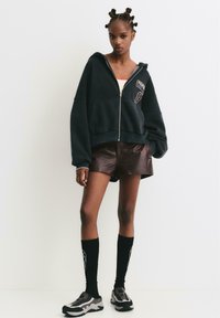 Čierna mikina na zips s logom, oversized strih, skombinovaná s hnedými koženými šortkami. Čierne ponožky po kolená a šedé športové topánky.