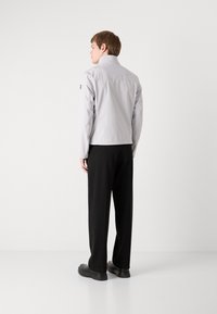 Ανοιχτό γκρι, ελαφρύ μπουφάν με όρθιο γιακά, που διαθέτει λογότυπο στο μανίκι. Συνδυάζεται με μαύρο παντελόνι σε χαλαρή γραμμή και μαύρα παπούτσια.