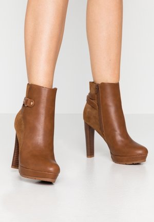 Kobieta nosząca brązowe skórzane botki na wysokim obcasie z bocznym zamkiem błyskawicznym i ozdobą z klamry, stojąca na białej powierzchni.