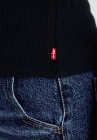 Fekete bordázott pulóver, az alján egy kis piros Levi's címkével, sötét farmernadrággal párosítva, amelynek narancssárga varrása van.