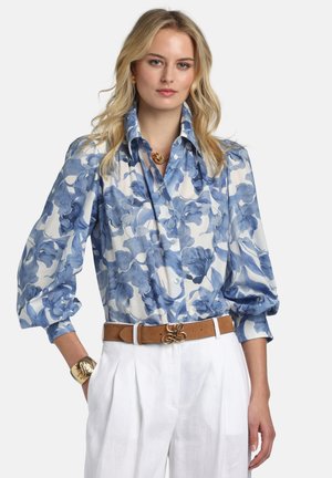 Blonde Frau trägt eine blaue und weiße Blumenbluse mit Puffärmeln, weiße Hose, braunen Gürtel, goldene Halskette, Ohrringe und Armband.