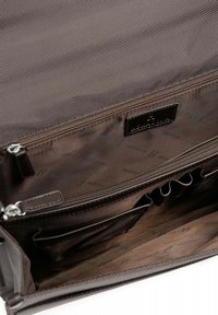 De binnenkant van een bruine stoffen tas met verschillende compartimenten, waaronder een ritsvak en insteekvakken, met een gladde textuur en logo-details.