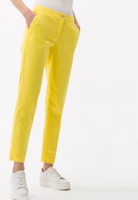 Pantalon en coton jaune à coupe slim, doté de poches latérales, d'une fermeture frontale à boutons et d'une texture lisse ; associé à des baskets blanches.