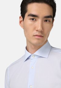 Lichtblauw gestreept overhemd met een klassieke kraag, voorzien van witte knopen en een gladde stoftextuur. Close-up van de bovenste torso van het model.