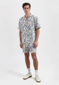 Korte mouwen, overhemd met kraag en bijpassende shorts in lichtblauw met een beige abstracte bladerenpatroon; de stof lijkt licht en gestructureerd.