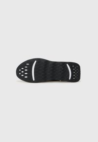 Semelle de chaussure en caoutchouc noir avec une surface texturée, présentant des découpes circulaires et rectangulaires, ainsi que des lignes d'accent blanches.