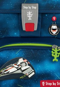 Uno zaino in tessuto blu presenta un design di navetta spaziale. Tra le caratteristiche ci sono un'etichetta grigia "Step by Step", una cerniera verde e un charm aggiuntivo.