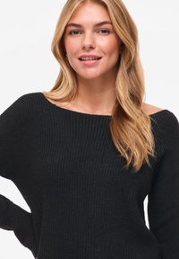 Μαύρο ριμπ πλεκτό πουλόβερ με σχεδιασμό έξω από τον ώμο. Διαθέτει μακριά μανίκια και υφή με ανάγλυφο τελείωμα. Το μοντέλο έχει κυματιστά ξανθά μαλλιά και ουδέτερη έκφραση.