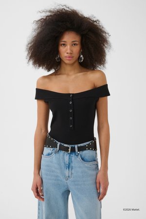 Woman with voluminous curly hair wearing off-shoulder black top with buttons, light blue jeans, studded black belt, and clear drop earrings.