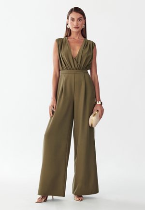 Kvinde iført ærmeløs olivengrøn jumpsuit med vide ben, holder en beige clutch, med langt, glat hår, guldbelagte øreringe og sandaler.