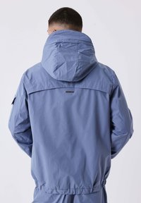 Uomo che indossa una giacca con cappuccio blu e pantaloni abbinati, visto da dietro su uno sfondo chiaro.