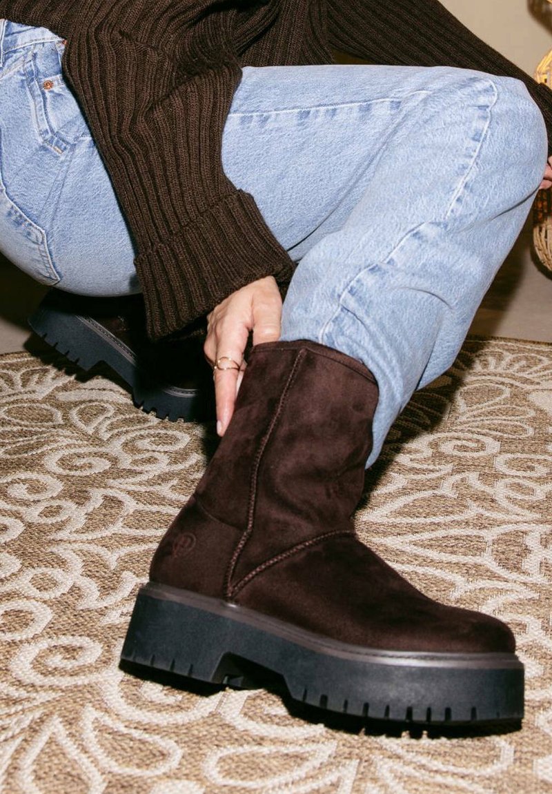 Stivali alla caviglia in suede marrone con una robusta suola in gomma nera, dotati di dettagli di cucitura discreti e di una finitura texture, posizionati su un tappeto a motivo.