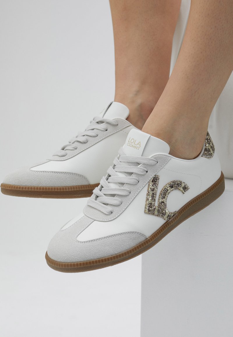 Zapatillas blancas con acentos de ante gris, un logo brillante "LC" en el lateral y una suela de goma. Presentan una puntera redonda y un diseño con cordones.