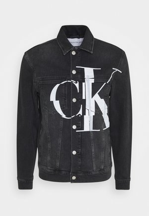 Schwarze Jeansjacke mit silbernen Knöpfen und großem, weiß überlappendem "CK"-Logo auf der Vorderseite, Kragen und sichtbaren langen Ärmeln.