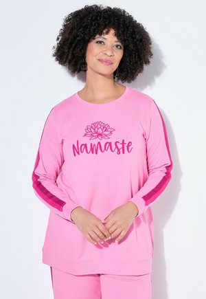 Kobieta o kręconych włosach ubrana w różowy top z długim rękawem i spodnie, na topie napis "Namaste" z motywem kwiatu lotosu nad tekstem.