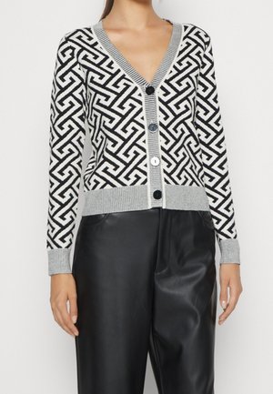 Femme portant un cardigan à motif géométrique noir et blanc avec bordure grise et grands boutons, associé à un pantalon en cuir noir.