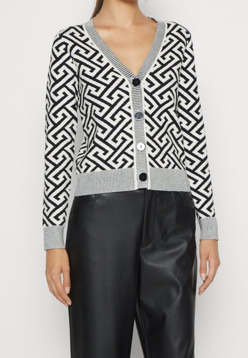 Femme portant un cardigan à motif géométrique noir et blanc avec bordure grise et grands boutons, associé à un pantalon en cuir noir.