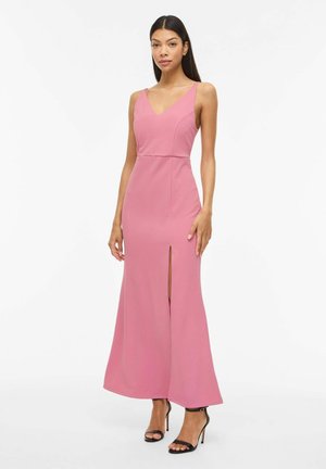 Frau steht, trägt ein ärmelloses pinkes Maxikleid mit Frontschlitz und schwarze Sandalen mit hohem Absatz, vor einem schlichten weißen Hintergrund.