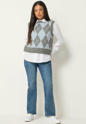 Femme debout portant une chemise blanche, un gilet en maille à losanges gris et bleu, un jean bleu et des baskets blanches sur un fond uni.