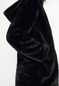 Cappotto in pelliccia sintetica nera con cappuccio, caratterizzato da una texture morbida, maniche lunghe e tasche laterali. La superficie ha un aspetto liscio e soffice.