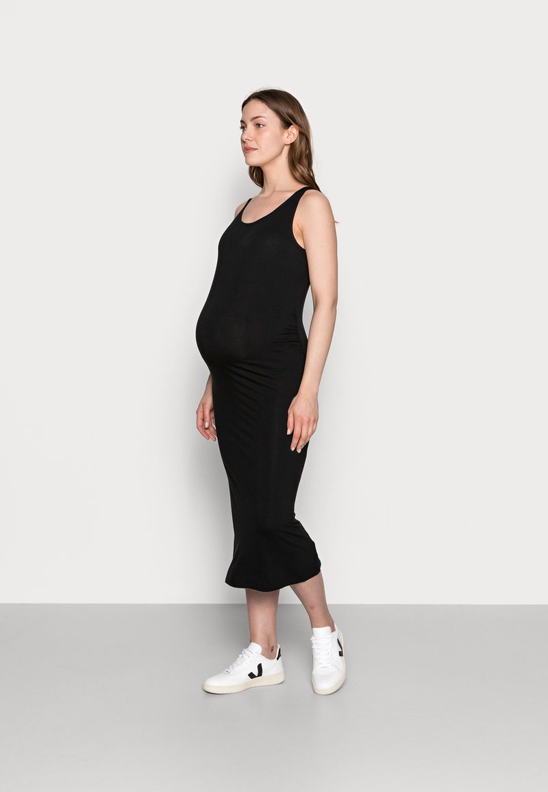 Vestido de maternidade preto com silhueta ajustada, decote redondo e bainha godê, combinado com tênis brancos com detalhes pretos.