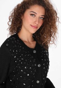 Czarny kardigan z dzianiny z okrągłym dekoltem, dekoracyjnymi czarnymi koralikami i srebrnym zdobieniem na przedzie.