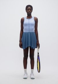 Γυναίκα με μπλε φόρεμα τένις κρατώντας ρακέτα, στέκεται μπροστά σε απλό λευκό φόντο, φορώντας λευκά αθλητικά παπούτσια και κάλτσες.