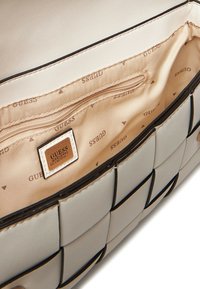 Interno di una borsa a mano beige intrecciata con una tasca con zip e etichetta con il logo "GUESS". Rivestito in tessuto color marrone chiaro con un motivo ripetuto del logo.
