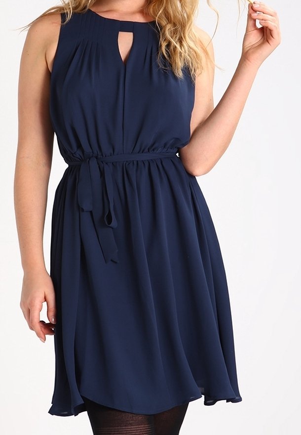 Femme portant une robe bleu marine sans manches avec un décolleté en œillet et une ceinture nouée à la taille, debout devant un fond neutre.