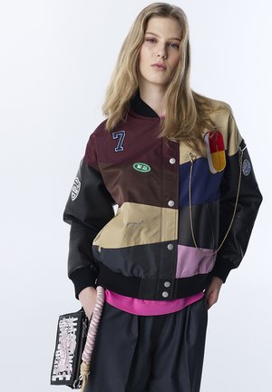 Jonge vrouw met blond haar draagt een felgekleurde patchwork bomberjack, zwarte broek en houdt een tas met een roze touwhandvat vast.