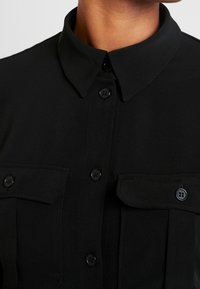 Acercamiento a una camisa negra de botones con cuello puntiagudo y dos bolsillos en el pecho con botones, utilizada por una persona.