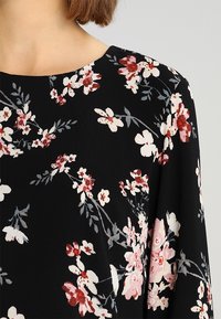Blouse noire à motifs floraux avec un col rond, ornée de fleurs blanches et rouges et de feuilles vertes, tissu texturé avec un tombé souple.