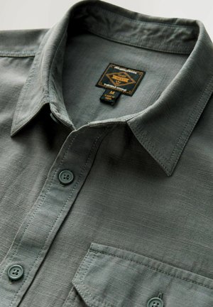 Sivina srajca z gumbi in ovratnikom, ki ima prsni žep, diskretno teksturo in oznako logotipa, pritrjeno znotraj ovratnika.