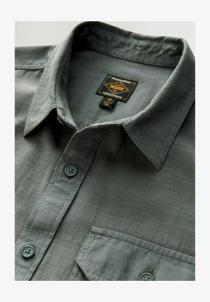 Pilkos spalvos marškinių su apykakle, turinčių krūtinės kišenę, subtilią tekstūrą ir logotipo etiketę, prisegtą vidinėje apykaklės pusėje.