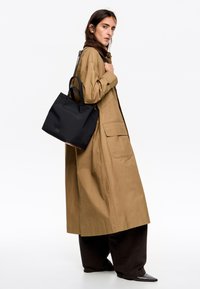 Zwarte draagtas met dubbele handvaten, gladde stof en logo-detail; gedragen met een tan jas; model poseert zijwaarts tegen een neutrale achtergrond.