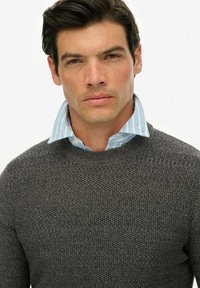 Szary sweter z dzianiny o teksturowanym wzorze, nałożony na jasnoniebieską i białą koszulę w prążki z kołnierzykiem. Prosty okrągły dekolt.