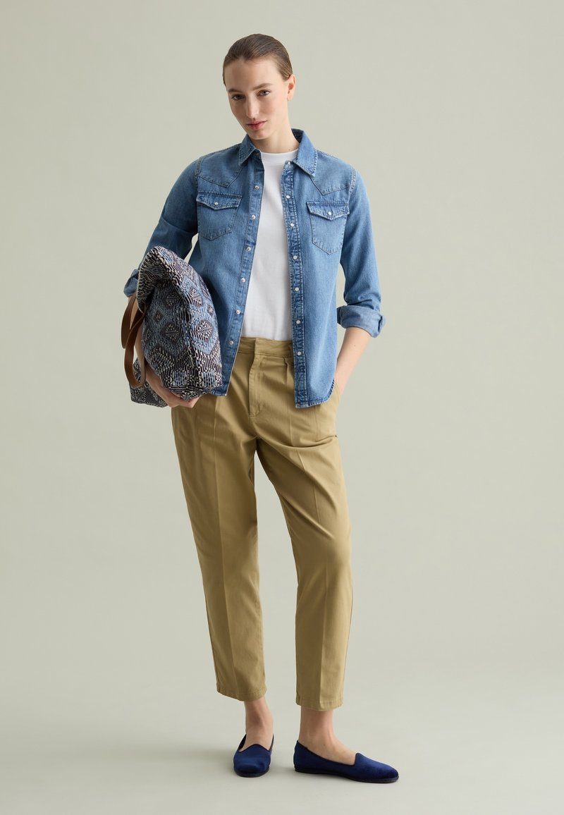Chemise en denim avec deux poches poitrine, T-shirt blanc, pantalon beige et ballerines navy. Tenant un sac à motifs avec des poignées marron.