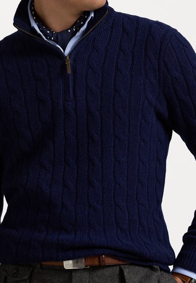 Navyblauwe kabelgebreide trui met een halve ritssluiting, met textuurpatronen en een zachte afwerking. Draag het over een lichtblauw overhemd met stippen.