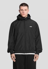 Model stoi na szarym tle, ubrany w czarną kurtkę z zamkiem błyskawicznym i kapturem, z białym logo, eksponując tatuaże na dłoniach.