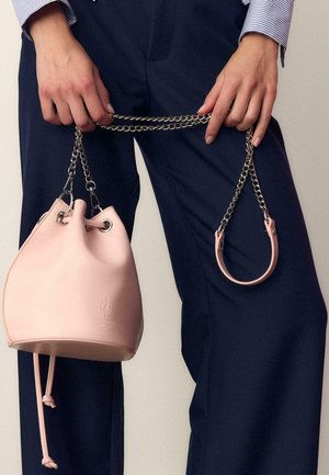 Person hält eine kleine hellrosa Bucket-Bag mit Kettenriemen, trägt marineblaue Hose und ein gestreiftes Hemd.